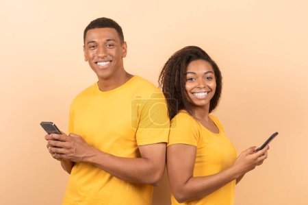 Téléchargez les photos : Happy african american couple with mobile phones posing on yellow studio background and smiling at camera. Black man and woman using cool mobile app on smartphones - en image libre de droit