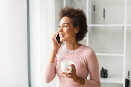 Photo for Surprised cheerful young mixed race lady calling by phone, enjoy communication and cup of coffee, look at copy space in living room interior. Emotions and reactions to good news and gossip at home - Royalty Free Image
