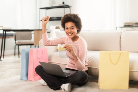 Téléchargez les photos : Satisfied young mixed race female shopaholic sit on floor with laptop, credit card, raising hand up celebrating success in room interior. Finance, banking, online shopping at home, sale and cashback - en image libre de droit