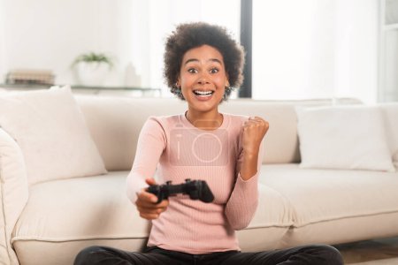 Foto de Cheerful excited young mixed race lady with joystick playing computer game, make victory and success gesture in living room interior. Win emotions, modern entertainment, fun alone at home with gadget - Imagen libre de derechos