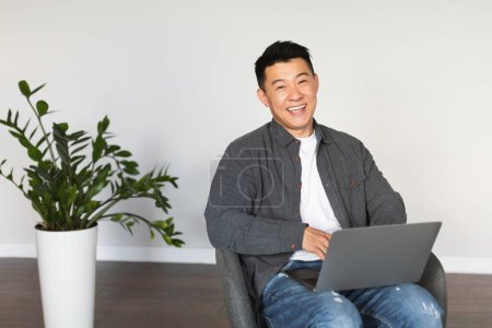 Téléchargez les photos : Smiling handsome mature asian guy sitting in armchair typing on computer, looking at camera, enjoy job in living room interior with white wall. Gadget for business, work, freelance remotely at home - en image libre de droit