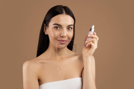 Téléchargez les photos : Smiling attractive good-looking half-naked millennial woman wrapped in bath towel holding small pink bottle, recommending nourishing anti-age face serum or oil for young skin, beige studio background - en image libre de droit