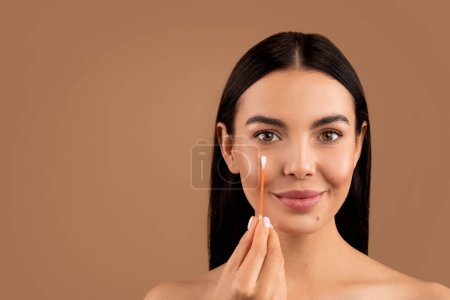 Photo for Beautiful brunette half-naked woman removes makeup from the face with a cotton swab. Happy young lady cleaning skin by cotton swab, beige studio background, headshot, copy space - Royalty Free Image