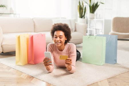 Photo for Cheerful young mixed race lady shopaholic with phone, credit card checking financial account lies on floor in living room interior with many packages from store. App for online shopping at home, sale - Royalty Free Image