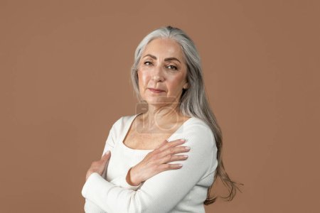 Téléchargez les photos : Serious pretty retired european female with gray hair looking at camera, isolated on brown background, studio, free space. Beauty and skin care, daily treatments, lifestyle and menopause, ad and offer - en image libre de droit