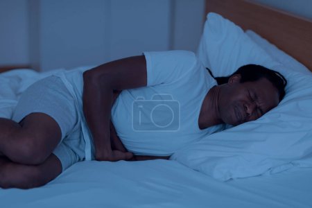 Foto de Nocturnal Panic Attack. Stressed Black Man Lying In Bed In The Night And Feeling Fear, Frustrated Young African American Man Awaken From Sleep, Embracing Himself And Frowning, Closeup - Imagen libre de derechos