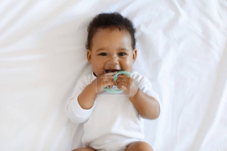 Photo for Portrait Of Adorable African American Baby Biting Teether And Looking At Camera While Lying On Bed At Home, Cute Black Infant Child Laughing And Playing With Toy While Relaxing In Bedroom, Top View - Royalty Free Image
