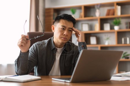 Téléchargez les photos : Unhappy mature asian guy analyzes data on laptop, presses hand to head, suffers from problems in business and headache in office interior. Error in work with device, stress, pressure and deadline - en image libre de droit