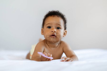 Foto de Childcare Concept. Portrait Of Cute Little Black Baby Lying On Bed With Teether In Hand, Adorable African American Infant Boy Wearing Diaper Looking At Camera With Interest While Resting At Home - Imagen libre de derechos