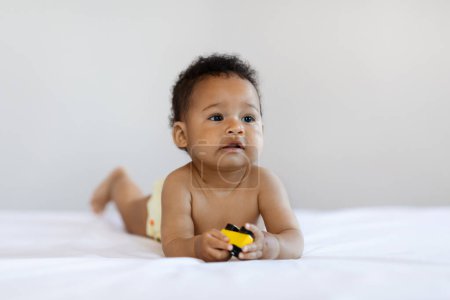 Foto de Portrait Of Adorable Black Infant Boy Playing With Toy Car While Lying In Bed On His Tummy, Closeup Shot Of Cute African American Baby Wearing Diaper Resting In Bedroom At Home, Copy Space - Imagen libre de derechos