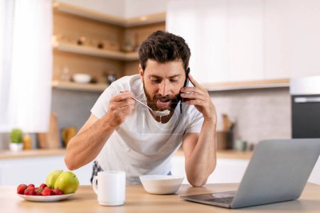 Téléchargez les photos : Busy cheerful married millennial caucasian guy with beard eats porridge, calls by phone, enjoys good morning, has breakfast in kitchen interior. Multitasking, work at home, communication and news - en image libre de droit
