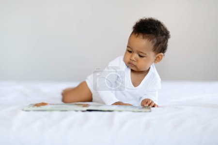 Foto de Baby Development Activities. Cute Little Black Boy Relaxing In Bed With Book, Curious African American Infant Child Looking At Pictures With Interest, Resting In Bedroom At Home, Copy Space - Imagen libre de derechos