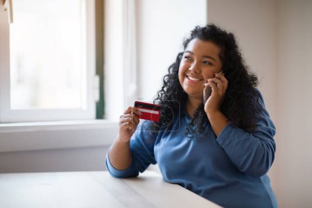 Photo for Pretty curly hispanic chubby woman looking at number on credit card and confirm purchase via telephone call to customer service, making payment via smartphone conversation, home interior, copy space - Royalty Free Image