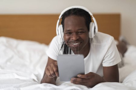 Téléchargez les photos : Happy Black Man With Digital Tablet And Wireless Headphones Relaxing In Bed, Cheerful Young African American Guy Using Tab Computer For Leisure At Home, Enjoying Modern Technologies, Closeup - en image libre de droit