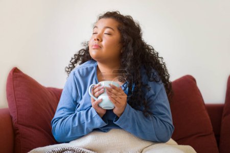 Foto de Delighted chubby mixed race curly young woman with a blanket on her lap sitting on the couch, holding mug, drinking hot tea with closed eyes, getting warm at home during cold winter, copy space - Imagen libre de derechos