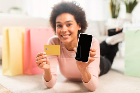 Foto de App for online shopping at home. Glad young mixed race lady shopaholic shows phone and credit card, lies on floor in living room interior with many packages from store. Sale, cashback and website - Imagen libre de derechos