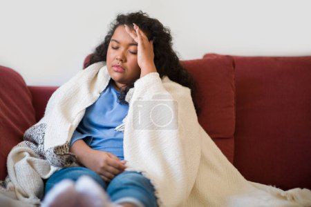 Foto de Sick unhappy plus size brunette curly young woman covered in warm blanket sitting on couch at home, touching head, suffering from headache during coronavirus or flu, copy space - Imagen libre de derechos