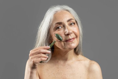 Téléchargez les photos : Happy elderly caucasian woman with gray hair doing jade massage to face with quartz roller, isolated on gray background, studio. Beauty care, anti-aging treatment, anti-wrinkle procedure, ad and offer - en image libre de droit