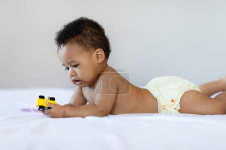 Téléchargez les photos : Beautiful Black Newborn Baby Playing With Toy While Relaxing On Bed At Home, Side View Of Cute African American Infant Child In Reusable Diaper Resting On His Tummy In Bedroom, Copy Space - en image libre de droit