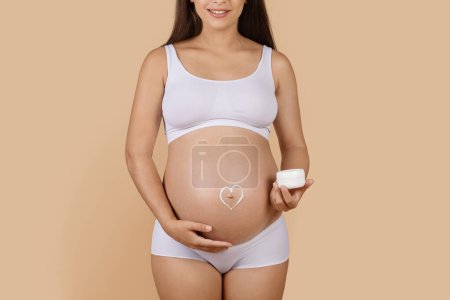 Téléchargez les photos : Pregnant woman with heart made of cream painted on her belly standing over beige studio background, cropped shot of expectant female in underwear applying moisturising body lotion, copy space - en image libre de droit