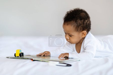 Foto de Adorable Black Baby Boy Or Girl Reading Book While Lying On Bed At Home, Cute Little African American Infant Child Making Development Activities While Resting In Bedroom, Closeup Shot With Copy Space - Imagen libre de derechos