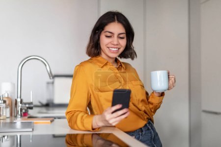 Téléchargez les photos : Online Communication. Portrait Of Beautiful Young Arab Woman Using Smartphone And Drinking Morning Coffee In Kitchen, Happy Middle Eastern Female Messaging With Friends And Drinking Tea At Home - en image libre de droit