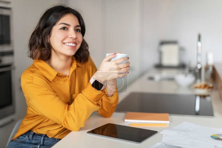 Téléchargez les photos : Smiling Young Arab Woman Drinking Coffee And Using Digital Tablet In Kitchen, Happy Middle Eastern Female Freelancer Sitting At Desk At Home, Enjoying Hot Drink And Looking Away, Free Space - en image libre de droit