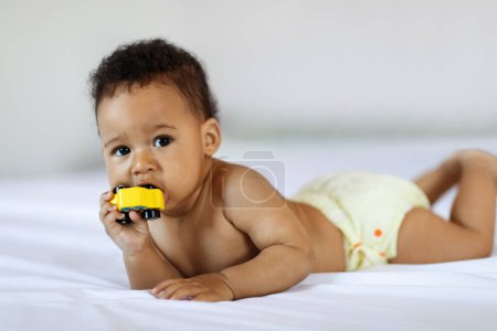 Foto de Teething Concept. Adorable Little Black Baby Biting Toy While Lying In Bed, Portrait Of Cute Infant African American Boy Or Girl Scratching Gums With Wooden Car While Relaxing In Bedroom, Copy Space - Imagen libre de derechos