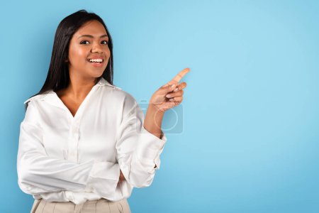 Téléchargez les photos : Smiling millennial black woman in white blouse pointing finger at free space, isolated on blue background, studio. Professional recommendation for business solution, work, study, startup, ad and offer - en image libre de droit