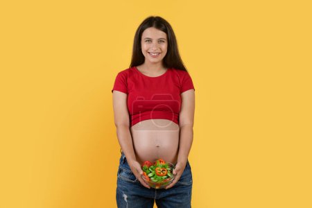 Téléchargez les photos : Beautiful Young Pregnant Woman With Bowl Of Fresh Vegetable Salad In Hands Standing Isolated On Yellow Background, Smiling Expectant Female Enjoying Healthy Vitamin Food During Pregnancy, Copy Space - en image libre de droit