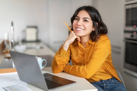 Photo for Dreamy Young Arab Female Sitting At Desk With Laptop In Kitchen, Happy Middle Eastern Woman Holding Pen And Looking Away, Daydreaming While Working Online With Computer At Home, Closeup - Royalty Free Image