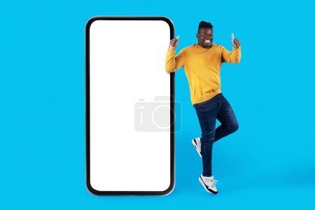 Téléchargez les photos : Great Offer. Happy Black Man Jumping Near Big Blank Smartphone With White Screen And Gesturing Thumb Up, Excited African American Guy Recommending Mobile App Or Website Over Blue Background, Mockup - en image libre de droit