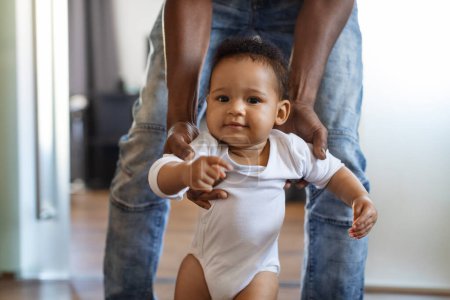 Téléchargez les photos : Closeup Of Adorable Black Infant Boy Walking With Fathers Help In Room, Cute African American Toddler Child Wearing Bodysuit Making First Steps At Home, Dad Supporting Him, Cropped Shot - en image libre de droit