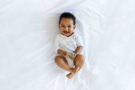 Foto de Child Care Concept. Top View Of Adorable Black Infant Boy Relaxing On Bed At Home, Cute Little African American Baby Wearing Bodysuit Holding Teether In Hand And Smiling At Camera, Copy Space - Imagen libre de derechos