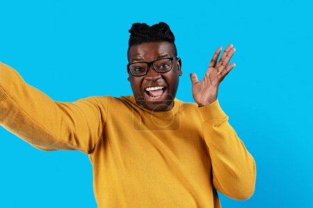 Foto de Cheerful Black Young Man Wearing Eyeglasses Taking Selfie Over Blue Background, Happy Excited Millennial African American Guy Having Fun While Making Self-Portrait, Looking And Smiling At Camera - Imagen libre de derechos