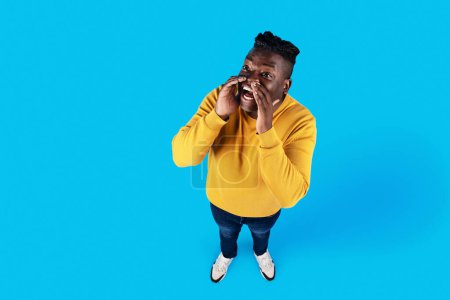 Téléchargez les photos : Cheerful Young Black Man With Hand Near Mouth Making Announcement, Above Shot Of Funny African American Guy Sharing News Or Information While Standing Isolated On Blue Background, Copy Space - en image libre de droit