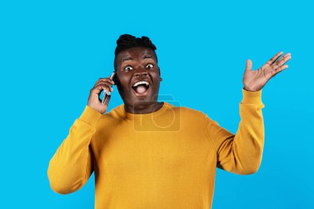 Téléchargez les photos : Mobile Call. Portrait Of Joyful Black Man Talking On Cellphone And Exclaiming With Excitement, Cheerful African American Male Enjoying Pleasant Phone Conversation While Standing Over Blue Background - en image libre de droit