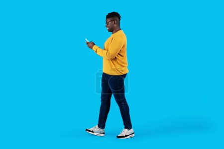Téléchargez les photos : Young Black Man Walking With Smartphone In Hands Over Blue Studio Background, Happy African American Male Messaging With Friends On Mobile Phone Or Shopping Online, Side View With Copy Space - en image libre de droit