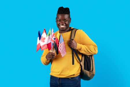 Photo for Study Abroad. Happy Black Man Holding Stack Of International Flags And Backpack, Smiling African American Male Student Enjoying International Education, Standing Over Blue Background, Copy Space - Royalty Free Image