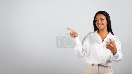 Foto de Happy pretty millennial black female in white blouse typing on smartphone, showing finger on free space isolated on gray background. Ad and offer, gadget for business, app for work, study and startup - Imagen libre de derechos