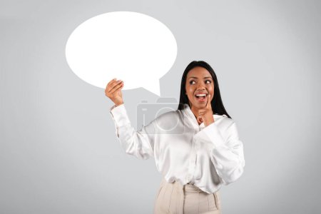 Téléchargez les photos : Laughing surprised millennial black female in white blouse with open mouth hold abstract bubble, cloud for words and thoughts isolated on gray background, studio. Idea for business, work, ad and offer - en image libre de droit
