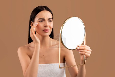 Foto de Stressed sad half-naked brunette young woman wrapped in bath towel looking at mirror and touching her face over beige studio background, having skin problems. Acne, pimples, wrinkles, dull skin - Imagen libre de derechos