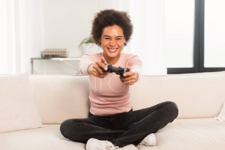 Foto de Cheerful excited young mixed race woman sitting on sofa with joystick, playing computer game in living room interior. Facial expression, entertainment, fun alone at home, gemer lifestyle and winning - Imagen libre de derechos
