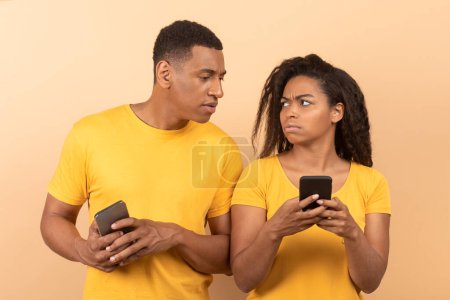 Foto de African american guy with smartphone spying on his wife chatting on mobile phone, yellow studio background. Black man does not trust his woman. Technologies and relationships concept - Imagen libre de derechos