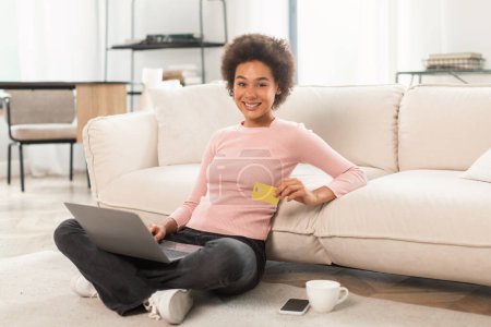 Téléchargez les photos : Happy young mixed race lady customer shopaholic sitting on floor with laptop and credit card in living room interior. Work, business, finance, banking and online shopping at home, sale and cashback - en image libre de droit