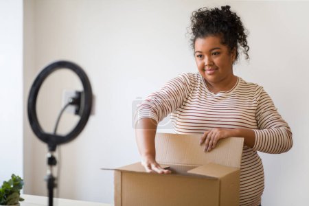 Photo for Cheerful pretty hispanic chubby millennial lady in casual outfit streaming while opening delivery parcel, standing next to paper box, smiling at smartphone on blogger set, home interior - Royalty Free Image