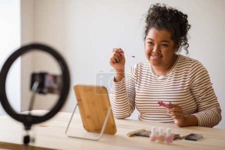 Téléchargez les photos : Cute young mixed race woman plus size beauty blogger sitting at table in front of mirror, recording video at home while applying lipstick, using phone, famous influencer recommending makeup products - en image libre de droit