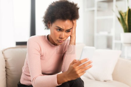 Foto de Frustrated unhappy young mixed race woman looking at smartphone, presses hand to head, suffering from migraine, depression and bad news in living room interior. Stress, problems, emotions at home - Imagen libre de derechos