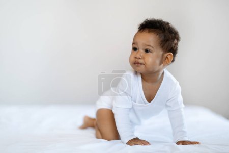 Foto de Child Care. Cute Black Baby Boy Or Girl Crawling On Bed At Home And Looking Away, Portrait Of Adorable Little African American Infant Kid Wearing Bodysuit Resting In Bedroom, Copy Space - Imagen libre de derechos