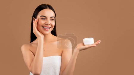 Téléchargez les photos : Attractive millennial long-haired brunette half-naked woman with perfect body and young smooth skin applying anti-aging face cream, looking at white jar on hand and smiling, beige studio background - en image libre de droit